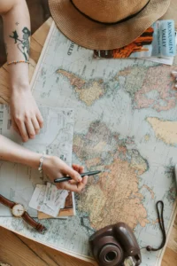 Photo d'illustration de l'article de blog Qu'est-ce que c'est un Travel planner ? qui montre la phase de préparation d’un voyage sur mesure. On y voit une carte du monde dépliée sur une table, autour de laquelle une personne trace des itinéraires à l’aide d’un stylo. Des billets d’avion, une montre et un guide de voyage complètent la scène, rappelant le processus d’organisation d’un projet de voyage. L’image illustre parfaitement le rôle du travel planner : analyser une destination, créer un itinéraire personnalisé et rassembler tous les éléments nécessaires pour que le voyageur parte serein. Elle transmet l’idée d’anticipation, de gain de temps et de liberté, valeurs essentielles de l’organisation de voyage à la Réunion et dans l’océan Indien.